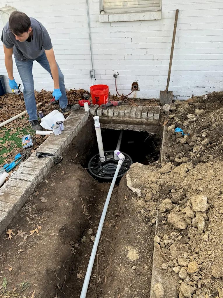 Technician performing exterior plumbing reroute and drain line repair beside a home foundation