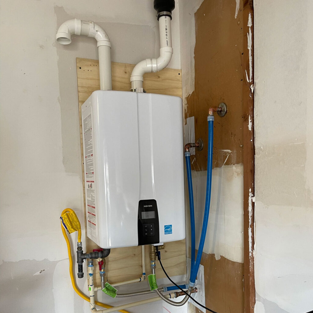 Wall-mounted tankless water heater with PVC venting, gas flex connector, isolation valves, and PEX lines installed on a backer board in a Fort Worth garage.