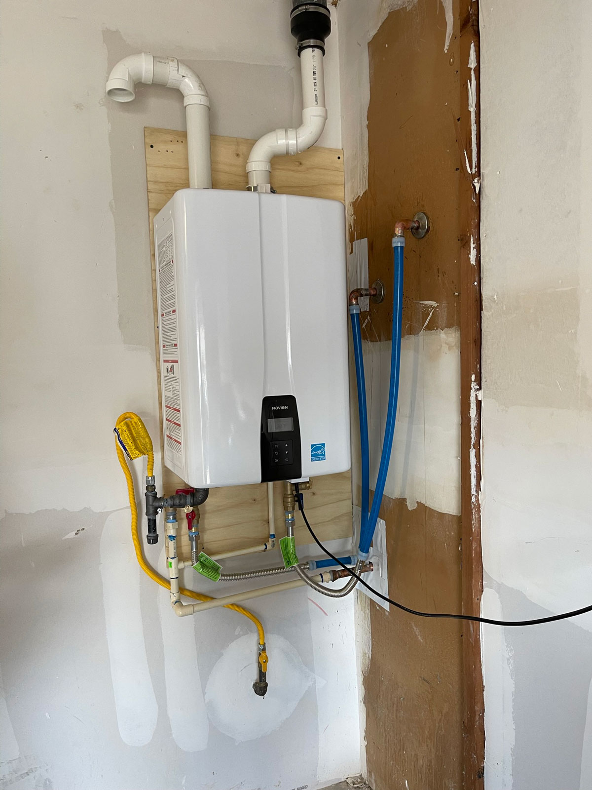 Wall-mounted tankless water heater with PVC venting, gas flex connector, isolation valves, and PEX lines installed on a backer board in a Fort Worth garage.
