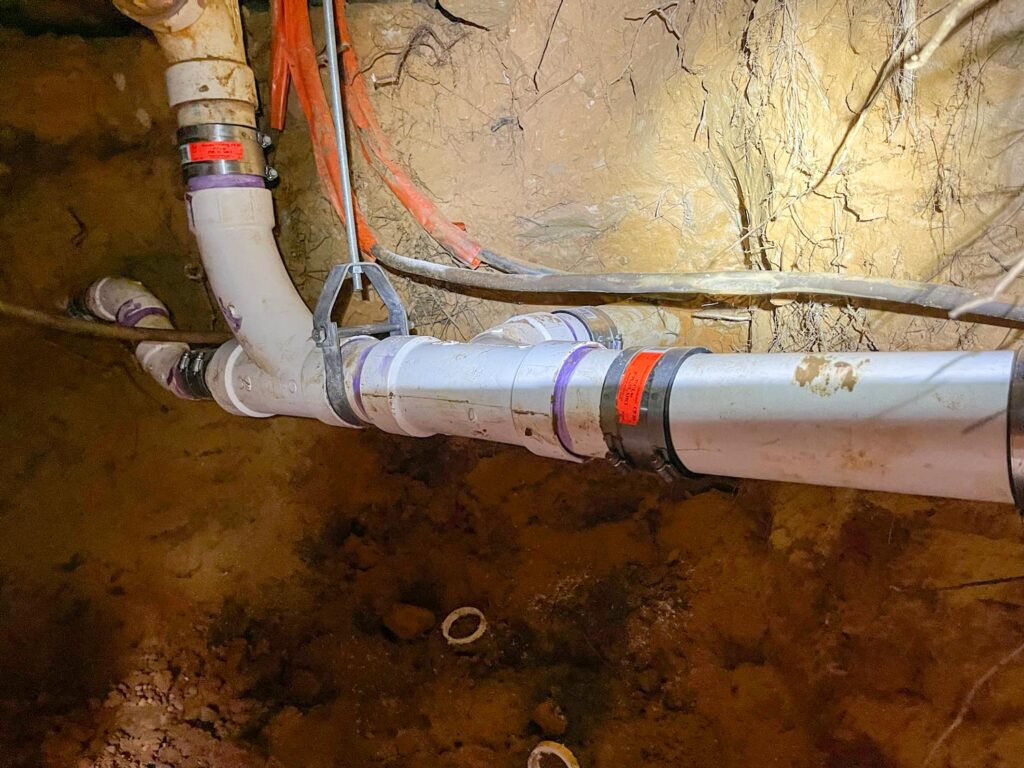 PVC drain line with shielded couplings inside a soil tunnel beneath a slab, showing clean primer joints and support straps during non-invasive tunneling repair.